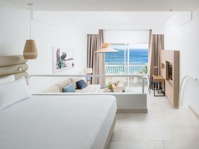 Helles, modernes Zimmer mit großem Bett, Sofa, TV und Meerblick durch bodentiefe Fenster.