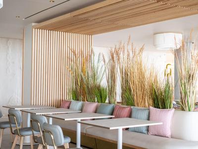 Moderner Essbereich mit langen Holztischen, grauen Stühlen und üppigen grünen Pflanzen im Hintergrund.