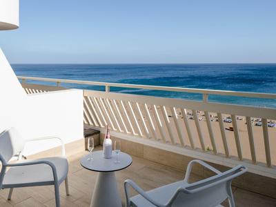 Balkon mit zwei weißen Stühlen, kleinem runden Tisch und Blick auf ruhiges, blaues Meer unter klarem Himmel.