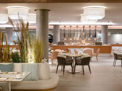 Moderner, heller Gastraum mit runden Tischen, Stühlen und dekorativen Pflanzen im Vordergrund.