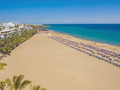 Ein Hotelpool mit Palmen, Liegestühlen und Blick auf einen breiten Sandstrand und ruhiges Meer.