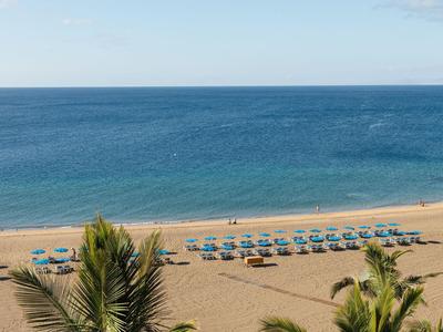 Strand met blauwe parasols en kalme zee achter palmbomen.