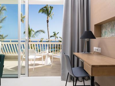 Heldere hotelkamer met bureau, stoel en balkon met uitzicht op zee en palmbomen.
