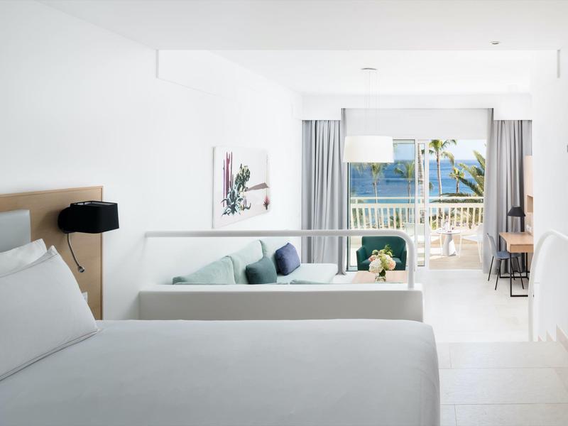 Hell eingerichtetes Hotelzimmer mit großem Bett, Sofa, Balkon und Meerblick bei Tageslicht.