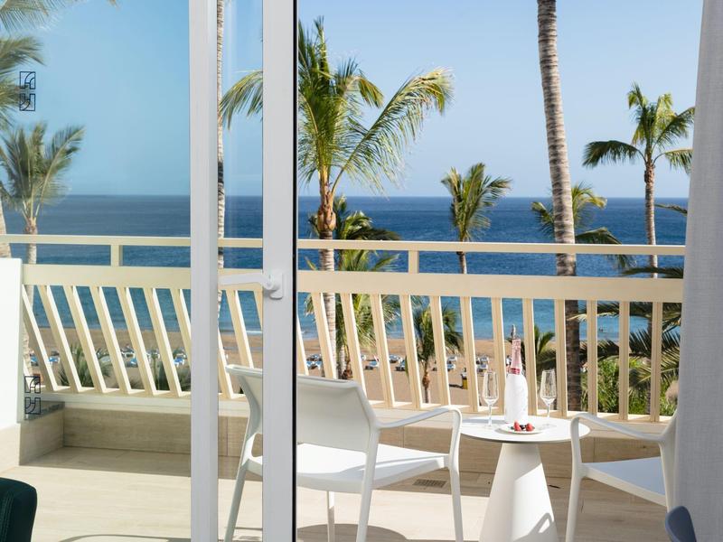 Balkon mit weißen Möbeln, Palmen und Blick auf das blaue Meer unter klarem Himmel.