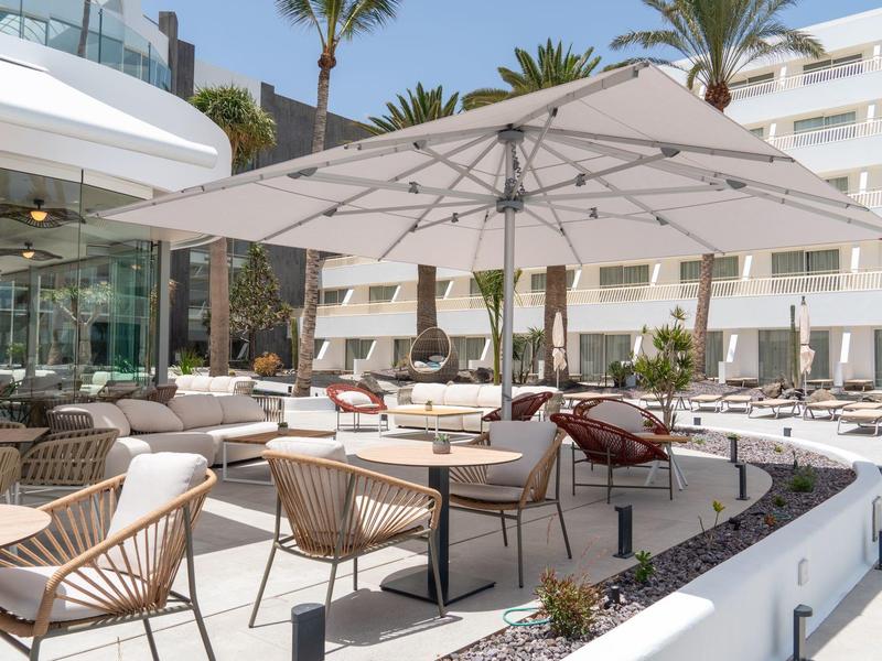 Moderne Außenlounge mit weißen Sonnenschirmen, Korbstühlen und Tischen unter Palmen.