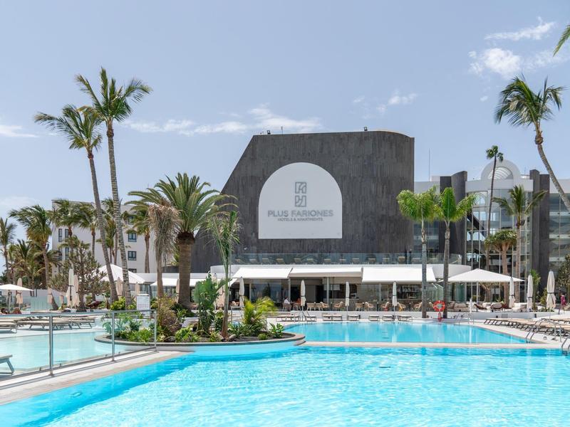 Großer Pool mit klarem Wasser, umgeben von Palmen und Liegestühlen vor modernem Gebäude.