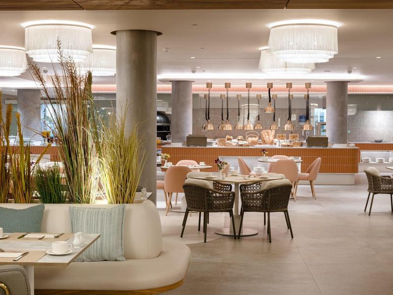 Moderner, heller Gastraum mit runden Tischen, Stühlen und dekorativen Pflanzen im Vordergrund.