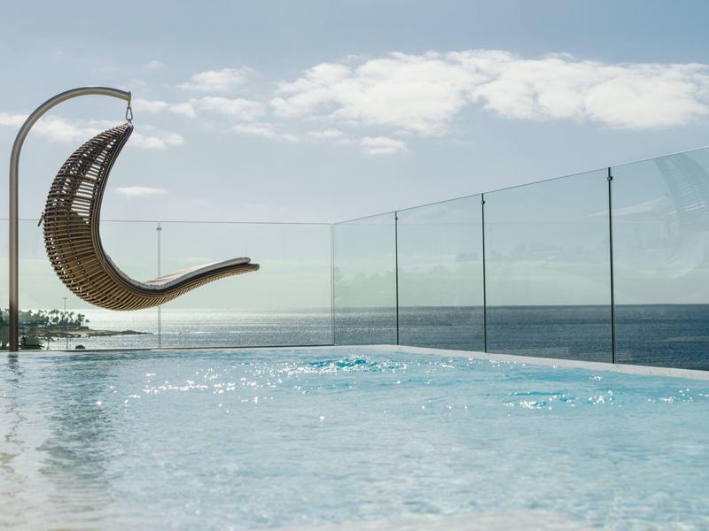Moderner Pool mit Glasgeländer und schwingendem Korbsessel vor Meer und blauem Himmel.