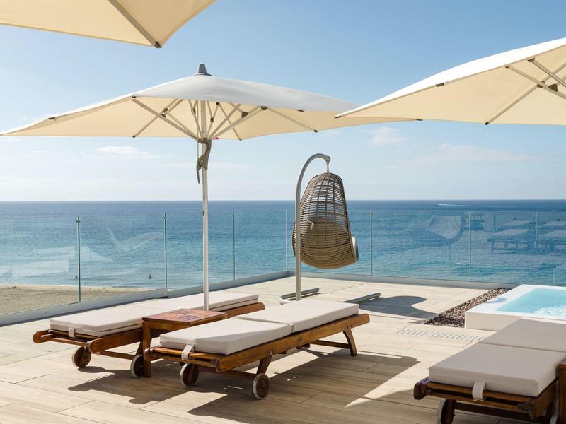 Terrassenbereich mit Liegestühlen, Sonnenschirmen und Blick auf das blaue Meer unter klarem Himmel.