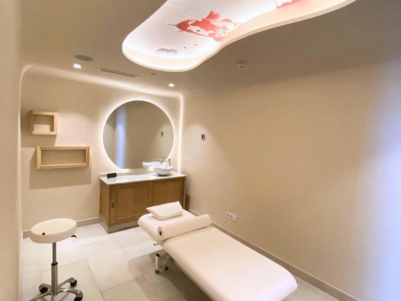 Moderner Behandlungsraum mit hellem Behandlungstisch, rundem Spiegel und bunter Deckenbeleuchtung.