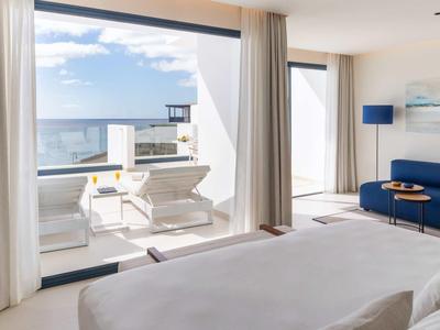 Modernes Hotelzimmer mit großem Bett, blauer Couch, Glaswand und Sonnenterrasse mit Meerblick.