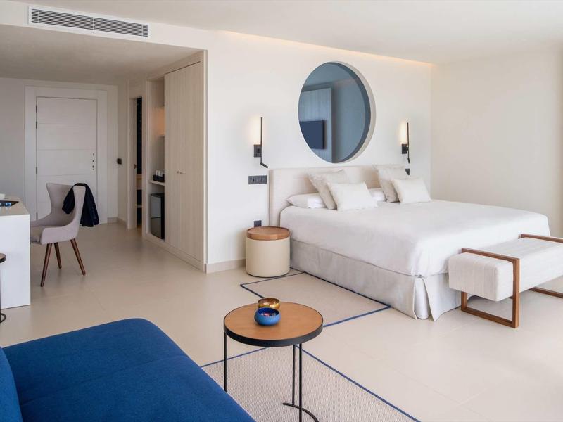 Helles, modernes Hotelzimmer mit Doppelbett, blauem Sofa, runden Spiegel und minimalistischem Mobiliar.