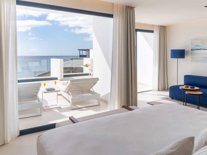 Modernes Hotelzimmer mit großem Bett, blauer Couch, Glaswand und Sonnenterrasse mit Meerblick.