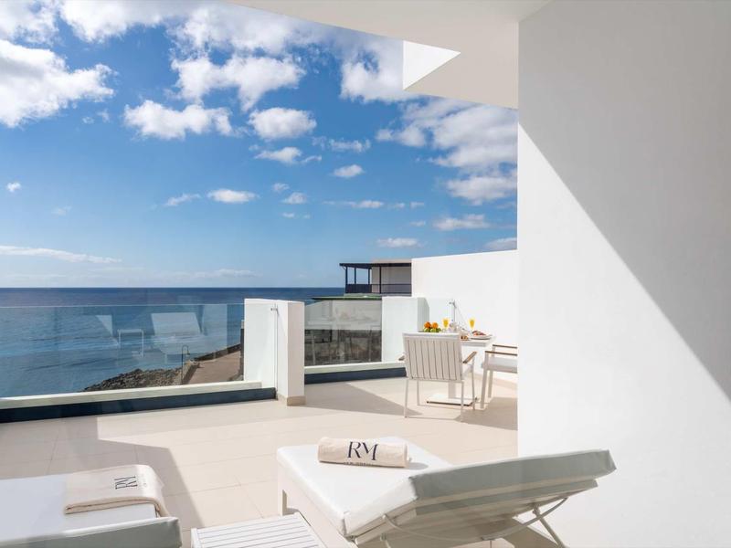 Moderne Terrasse mit Sonnenliege, Glasgeländer und Meerblick unter blauem Himmel mit Wolken.