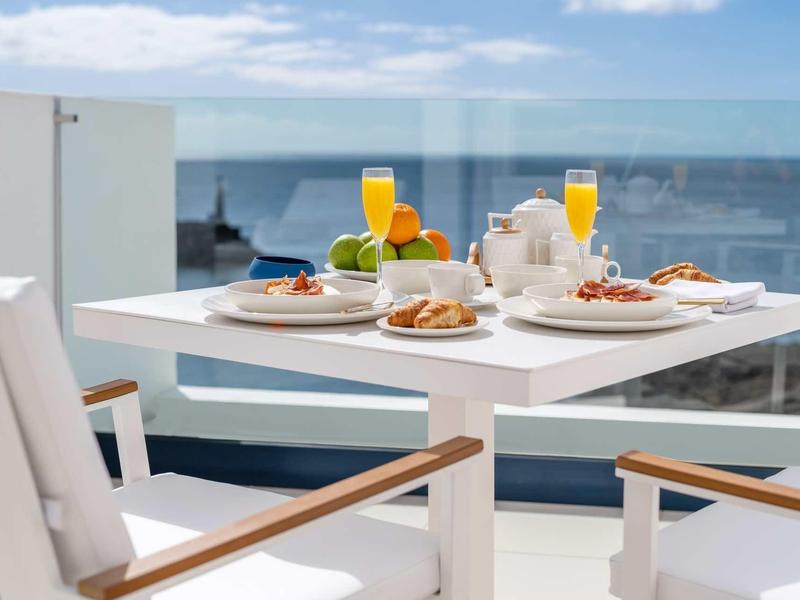 Weiß gedeckter Tisch mit Frühstück, zwei Orangensaftgläsern, auf Balkon mit Meerblick bei blauem Himmel.