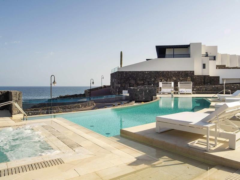 Moderner Außenpool mit Liegestühlen, klarem Wasser und Blick auf das Meer bei sonnigem Himmel.