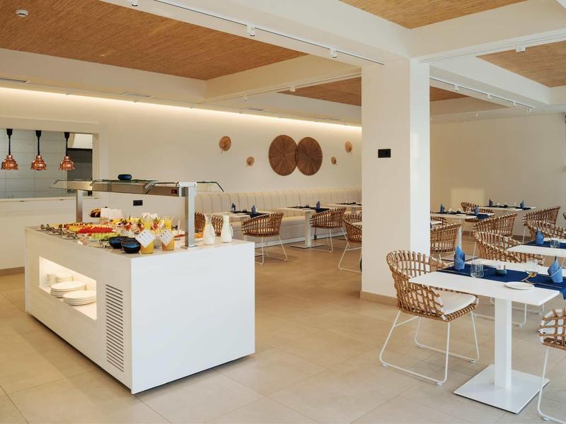 Moderner Speiseraum mit weißen Tischen, Stühlen aus Korbgeflecht und Buffetinsel in hellem Design.