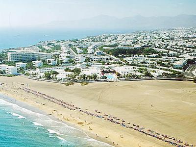 Langer Sandstrand mit blauem Meer, dahinter viele weiße Gebäude und grüne Vegetation, Berge im Hintergrund.