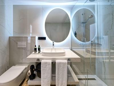 Nowoczesna łazienka z toaletą, umywalką, okrągłym podświetlanym lustrem i prysznicem z szklaną przegrodą.