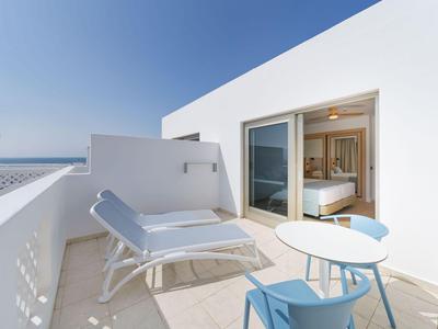 Jasny taras z leżakiem, niebieskim krzesłem i małym stolikiem przed oknem hotelowym z widokiem na morze.