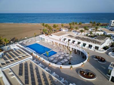 Vue d'un hôtel moderne avec piscine, terrasses ensoleillées et plage en arrière-plan.