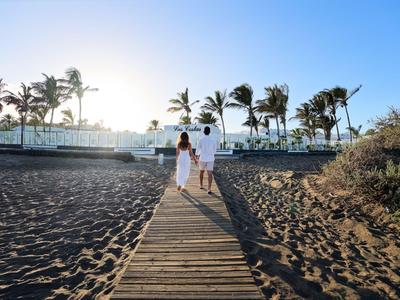 Un couple marche sur une passerelle en bois le long de la plage avec des palmiers et des bâtiments en arrière-plan.
