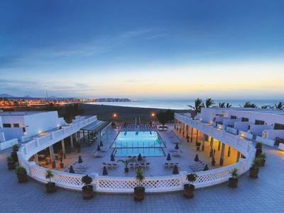 Moderne, weiße Hotelanlage mit Pool in der Mitte bei Sonnenuntergang am Meerufer.