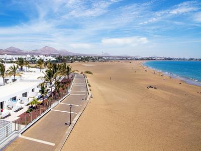 Breiter Sandstrand mit blauem Meer, Palmen, Promenade und weißen Gebäuden unter blauem Himmel.