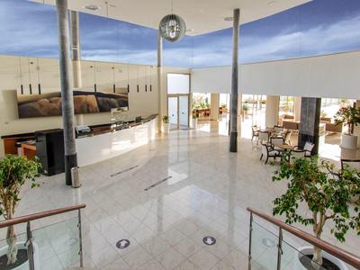 Heller, moderner Aufenthaltsraum mit weißen Fliesen, Sitzgruppen und großen Fenstern unter blauer Decke.