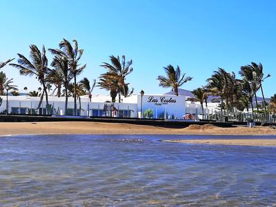 Strand mit ruhigem Wasser, dahinter weiße Gebäude und Palmen vor klarem blauem Himmel.