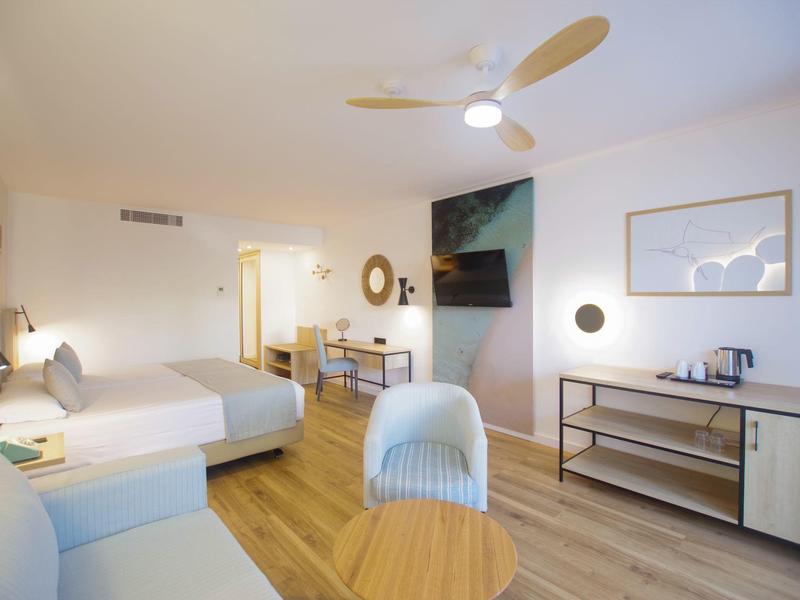 Moderner Hotelraum mit hellem Holzboden, großem Bett, Fernseher, Schreibtisch und Sitzbereich.