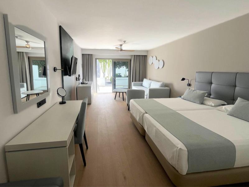 Helles Hotelzimmer mit zwei Betten, Schreibtisch, Sofa, Fernseher und Zugang zur Terrasse.