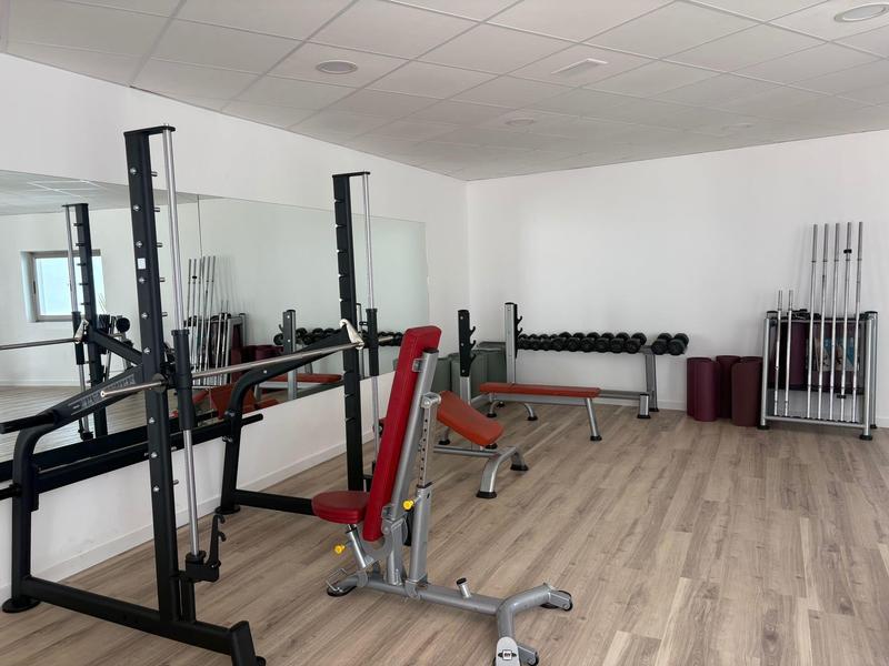 Moderner Fitnessraum mit Hantelbank, Kurzhanteln und Trainingsgeräten in hellem Raum.