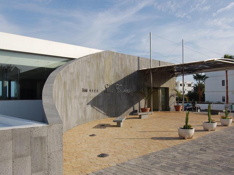 Moderner Gebäudeeingang mit geschwungener Betonwand, Glasfenstern und gepflastertem Platz.