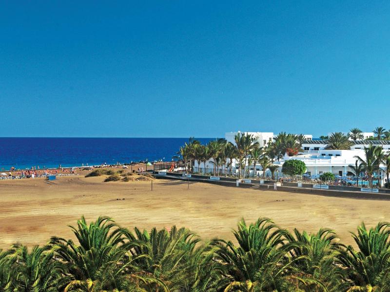 Strand mit goldfarbenem Sand, blauem Himmel, Palmen im Vordergrund und weißen Gebäuden am Rand.