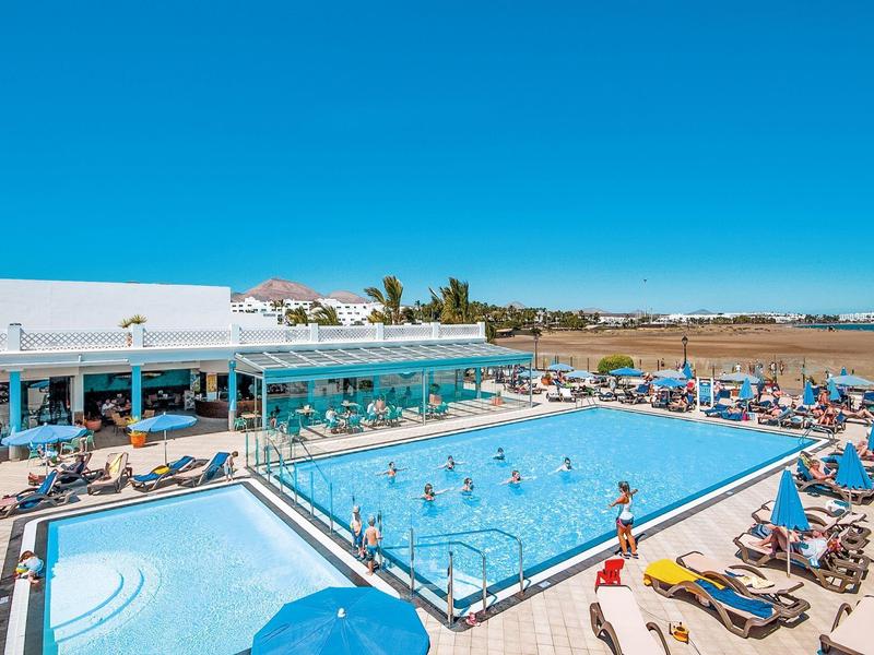 Schwimmbadbereich mit Rechteck- und Kinderbecken, vielen Sonnenschirmen, Liegestühlen und blauem Himmel.