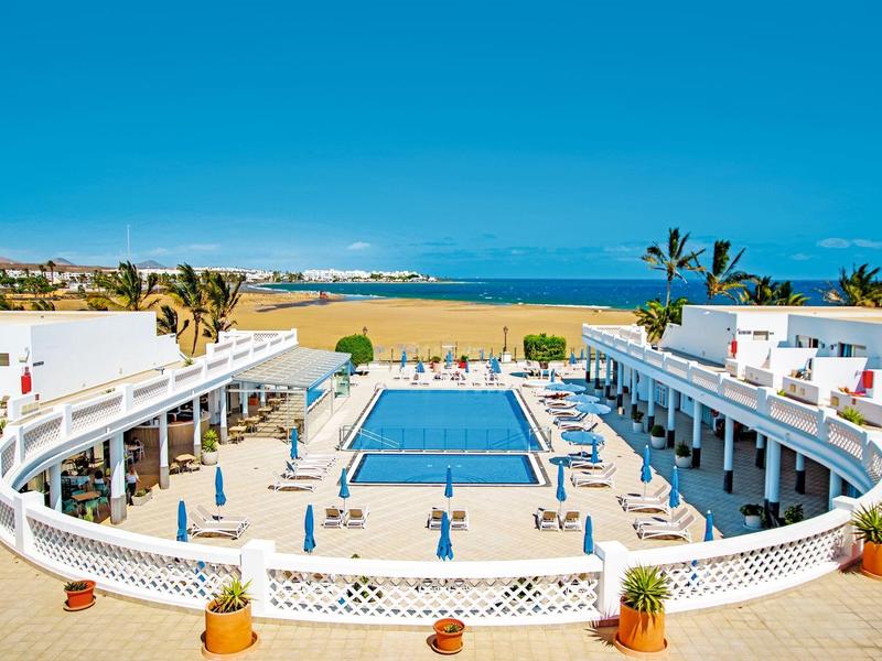 Weißes Hotel mit blauem Pool, Palmen und Blick auf goldenen Sandstrand und Meer im Hintergrund.