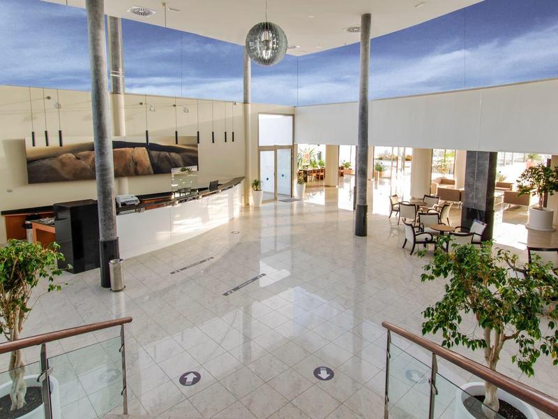 Heller, moderner Aufenthaltsraum mit weißen Fliesen, Sitzgruppen und großen Fenstern unter blauer Decke.