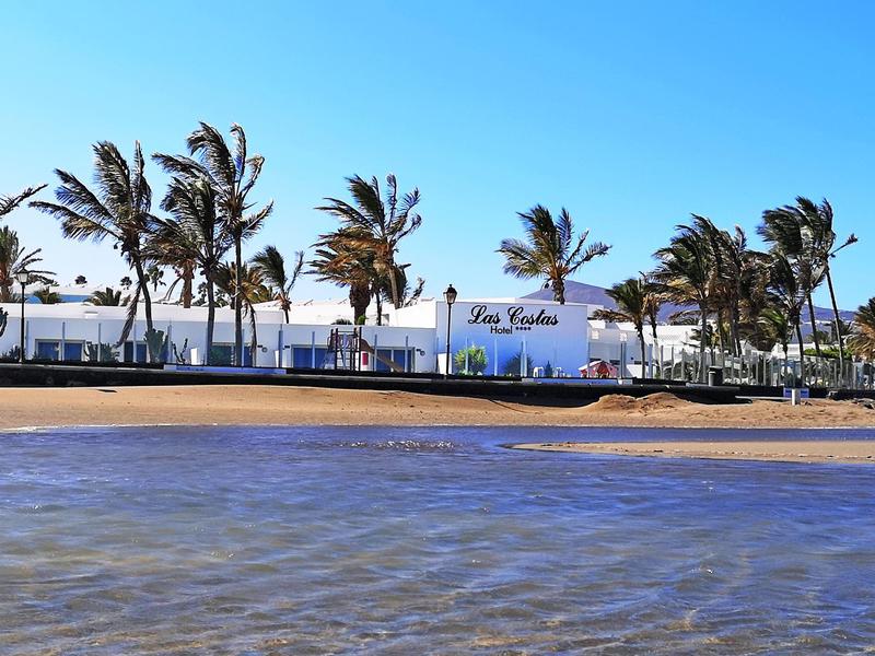 Strand mit ruhigem Wasser, dahinter weiße Gebäude und Palmen vor klarem blauem Himmel.