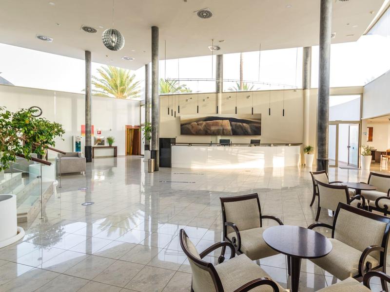 Helle und offene Hotellobby mit glänzendem Boden, Pflanzen, Sitzgruppen und Empfangstresen.