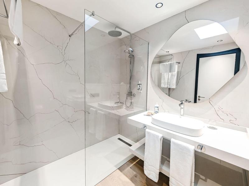 Modernes Badezimmer mit großer Glasdusche, weißem Waschbecken und rundem Spiegel. Boden hellbraun.