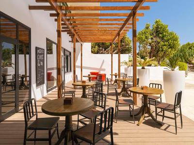 Außenbereich mit Holzterrasse, Tischen, Stühlen und Holzpergola neben Glasfenstern und grünen Pflanzen.