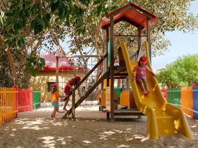 Kind spielt auf gelber Rutsche in buntem Spielplatz mit Baum und Sand unter dem Spielgerät.