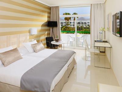 Helles Hotelzimmer mit Doppelbett, grauen Akzenten, Schreibtisch, Stuhl und Balkon mit Meerblick.