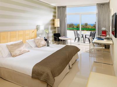 Hell eingerichtetes Hotelzimmer mit Doppelbett, Schreibtisch und Balkon mit Meerblick.