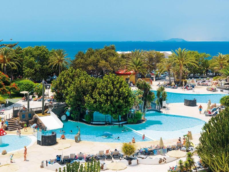 Großer Poolbereich mit Palmen, Liegestühlen und Blick auf das blaue Meer im Hintergrund.