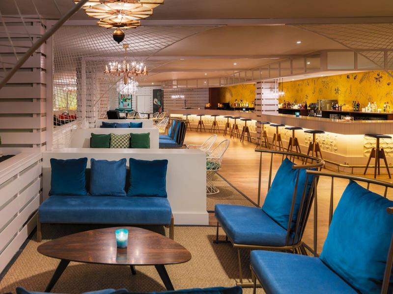 Moderner Lounge-Bereich mit blauen Sesseln, Holztischen und beleuchtetem Barbereich im Hintergrund.