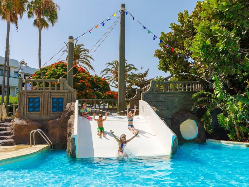 Kinder rutschen eine weiße Wasserrutsche in einem blauen Pool mit Palmen und grünen Bäumen.