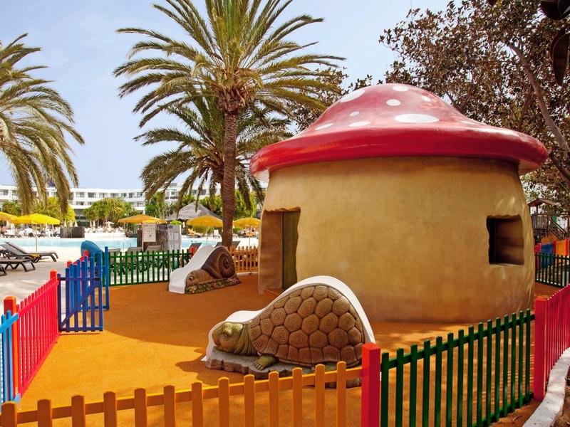 Bunt eingezäunter Spielplatz mit Pilzhaus, Palmen und Sandboden unter blauem Himmel.